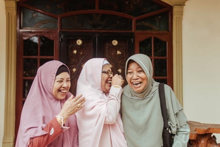 Three older Muslim women standing together and laughing, one in dark pink, another in light pink, and another in green hijabs.