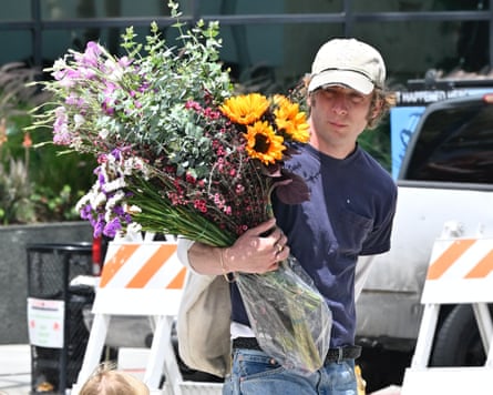 A photo of actor Jeremy Allen White wearing a beige baseball cap, navy T-shirt and jeans. He is carrying in his right arm a very large bunch of flowers, including sunflowers and irises. He is on a street that, in the background, has construction signs and a white car.