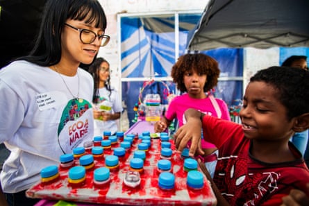 A young woman sits across from a smiling child who is opening bottles, watched by another child