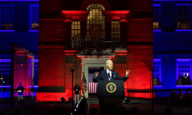 Joe Biden speaks at a podium emblazoned with the seal of the US presidency in front of a historic building bathed in dark red and navy blue lights.