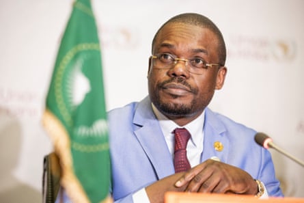 An African man in a pale blue suit at a desk on a podium in front of a mic with the green flag of the African Union in front of him