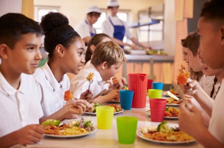 Primary school kids eat lunch in school cafeteria with brightly coloured water glasses.