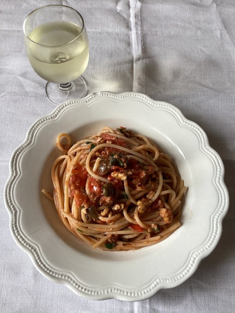 On a slightly wrinkled tablecloth sit a glass of white wine and a bowl with scalloped edges containing a pile of spaghetti swirled around with sauce on top