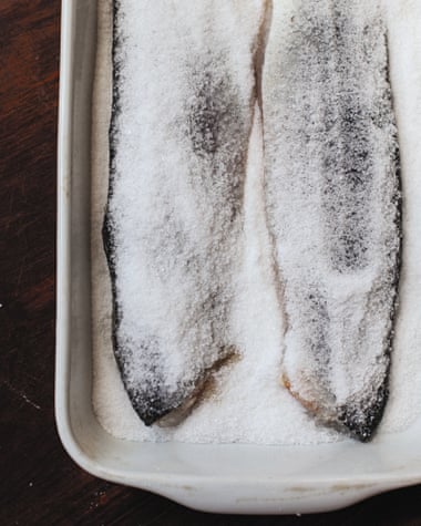 Due pezzi di filetti di branzino sono uno accanto all'altro in un piatto bianco intenso, seduti sopra il sale e lo zucchero che ricoprono anche i filetti.