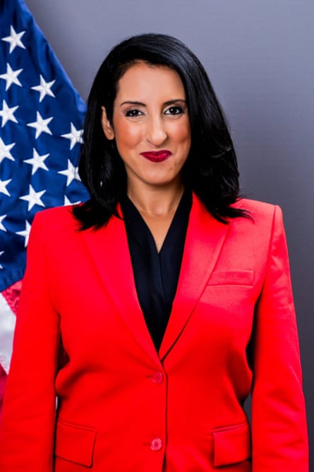A woman poses for a headshot wearing a bright red suit jacket and an American flag in the background
