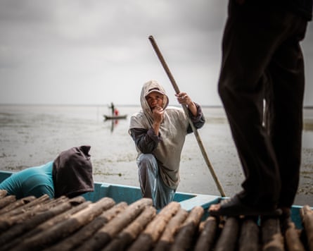 A man stands in a small boat holding a pole while another man stands on a jetty and another lifts something out of the boat