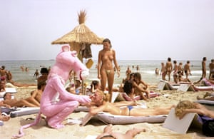 A person in a pink panther costume crouches next to a woman sunbathing topless on the beach, as another woman watches on