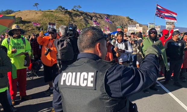 A police officer with his back to the camera addresses a group of people holding flags and signs.