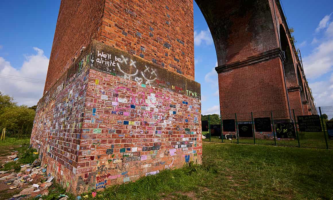 The Twemlow Viaduct in Holmes Chapel is photographed against a blue sky. the bright graffiti scrawled on it by Harry Styles fans visible. Just beyond the main section with graffiti, the protective fence erected by Network Rail can be seen