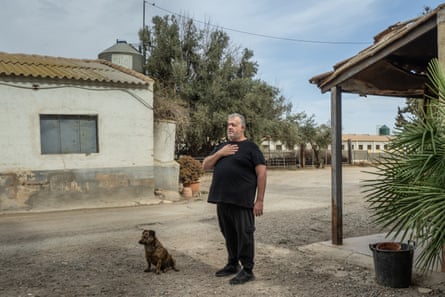 José está con su perro afuera de una casa, con cobertizos agrícolas apenas visibles en el fondo.