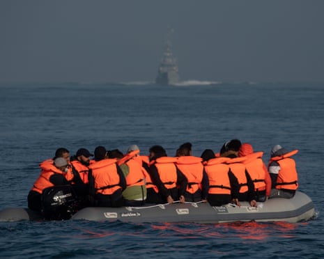 A crowded inflatable dinghy with people wearing orange life jackets floats on calm water in the Channel