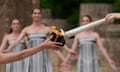 Close-up of the Olympic torch being lit from a bowl of flames as three women in dresses watch in the background