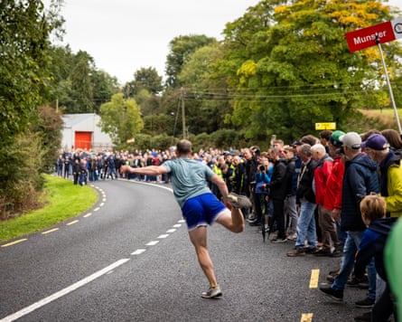 A man, his back to the camera, steps forward after hurling a ball down a road as spectators look on.