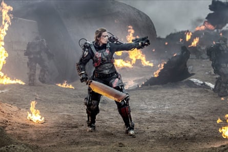 Emily Blunt wearing a mechanical battle suit, standing on a battlefield with patches of fire around her, holding some kind of weapon