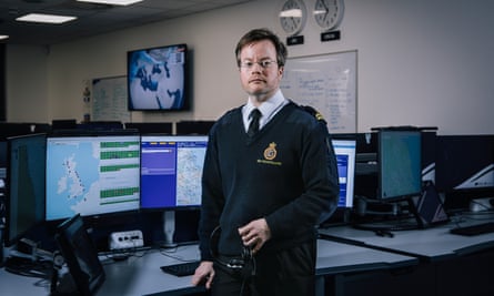 Coastguard Will Cook in Bridlington wearing uniform, holding headphones and standing by his desk with a large screen showing Great Britain, other screens around the office and two clocks on the wall.