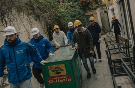 People in hard hats wheel out possessions from the pizzeria stored in a large green box
