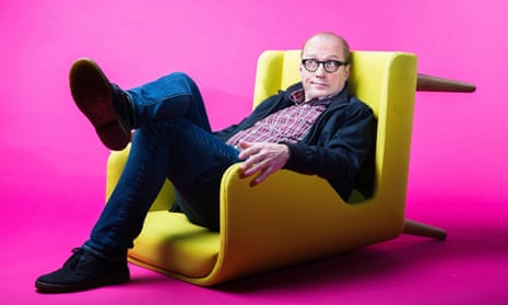 Comedian Adrian Edmondson sitting in upturned yellow chair against pink background