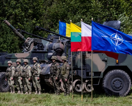 On grasslands, surrounded by trees, soldiers in the Polish and Romanian armies stand beside a number of flags, including the Nato flag, and military vehicles