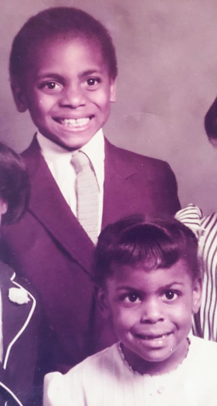 A crop of a sepia-tone image of a little boy in a suit smiling, with a little girl in a dress in front of him, and people obviously to their left and right.