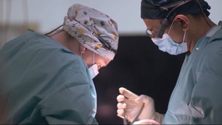 Two medics gowned up with hats and face masks look down as they perform an operation