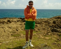Bad Bunny in an orange hoodie, yellow shirt, and green shorts stands with arms crossed by the ocean