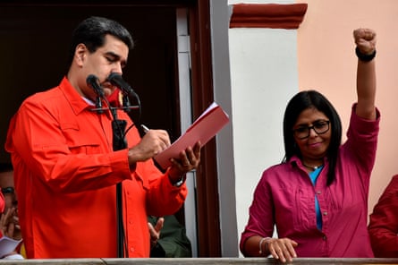 Delcy Rodríguez, wearing a fuschia shirt holds a clenched fist in the air as she stands next to Nicolás Maduro, wearing his signature red shirt, as he signs a document breaking off diplomatic ties with the US in Caracas in 2019