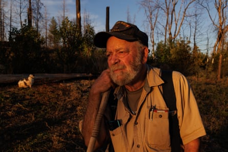 An elderly white man with a full gray beard, dark baseball cap, and large suspenders over a work shirt holds a shovel or other implement with his right hand and rests his chin against it.