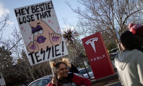 Protester with a placard that says 'Hey besties, lose that Tessie' next to Tesla branding