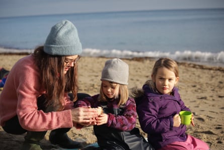Mother in woolly hat on a beach examines object with young daughter while another daughter holds a cup.