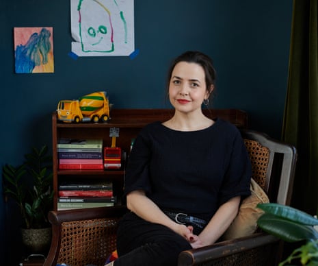 Joanna Wolfarth sitting at home, a yellow toy truck on a shelf and children's paintings on the wall behind her