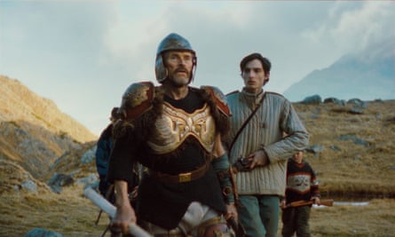 A young man and an older man, wearing armour and an armour helmet, in front of mountains