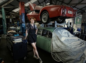 A women stands in a mechanic workshop next to two vintage cars.