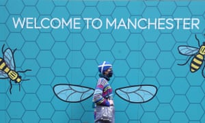 A person wearing a face mask walks past a Welcome to Manchester mural.