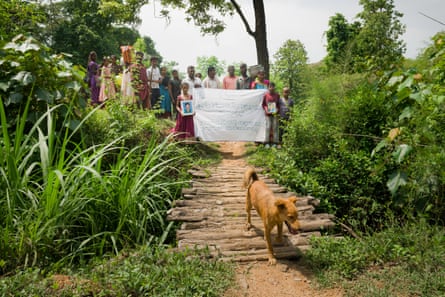 Sixteen or so Indians stand on a forest path holding a white sheet with writing in an Indian script and farmed photographs of a man and a woman