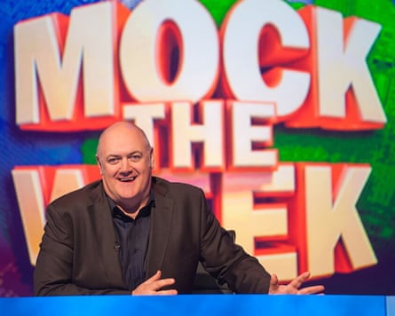 He sits at a desk in front of a large logo of the programme