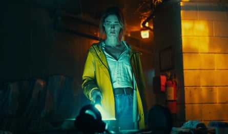A woman in a white blouse, jeans and a yellow rain jacket looks concerned as she shines a torch on a desk