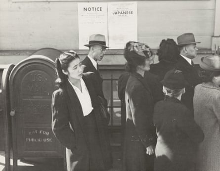 Black-and-white image of small crowd of well-dressed people, with the men and most of the women wearing hats, as one woman in the group steps out to apparently look up a street.