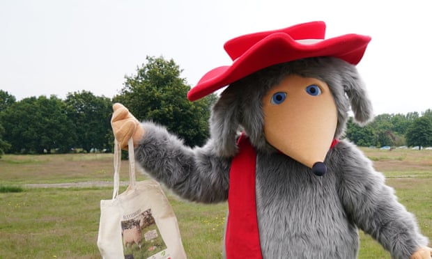 A Womble in a red heart and scarf standing in a field holding a canvas bag
