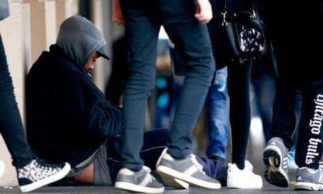 A homeless man sits on a street as people walk by in Auckland
