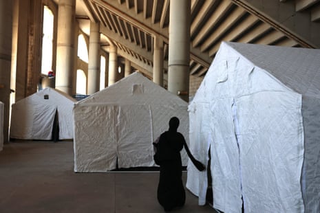 A woman dressed all in black and wearing a black hijab faces away from the camera as she enters a large white tent. Ine the background, there are two more large white tents, all standing in what appears to be the mezzanine level of a stadium.