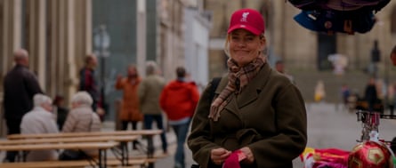 she is in her late 40s with blond hair and wears a red Liverpool Football Club baseball cap, a tartan scarf and khaki green coat. She is standing in a public space with benches and people in the background, with a rather wry smile on her face