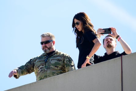 a woman stands on a roof as a man in camo gestures and a man behind her holds up his phone