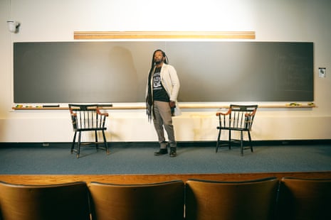 Black man with very long dreads, wearing black T-shirt, cream blazer, and gray khakis poses in front of long chalkboard between two chairs.