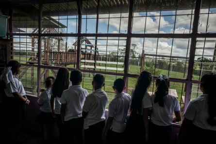 Nove crianças em uniforme escolar olham pela janela para um playground com uma plantação do outro lado