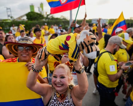 A smiling woman holds up a soft toy tiger dressed in a Colombia football top, as other supporters behind wave flags