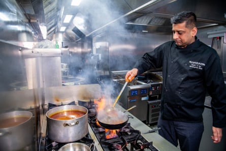 A man in a restaurant kitchen, stirring food in a metal bowl over an open flame, smiling for the camera