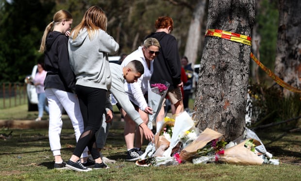 A group of mourners watch a young man lay a bouquet of flowers against a tree