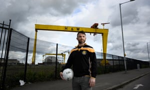 Dave McGreevy in a team shirt standing in front of two very large yellow cranes bearing the logo 'H&W'