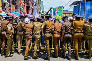 A row of police officers with their backs to the camera use yellow control barriers to hold back protesters holding up an image of the former president
