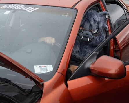 Chase Che, wearing a furry gray wolf mask, sits in the driver's seat of his orange Mazda6